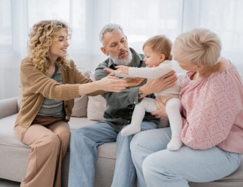 cheerful blonde woman outstretching hands to little daughter near parents on couch in living room